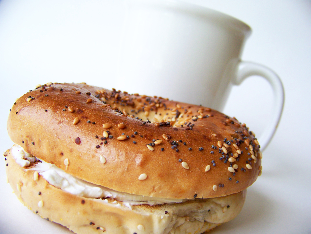 Bagel filled with cream cheese with a white coffee cup behind it. | Ellisville, MO